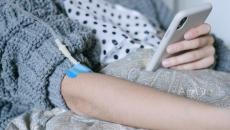 Patient in a blue sweater comforter recuperates at home and receives an IV infusion while checking patient portal information on a cell phone.