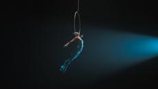 Trapeze artist in a blue costume holding onto a hoop in mid-air against a black backdrop