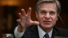 FBI Director Christopher Wray reaches his hand forward as he speaks into a microphone.